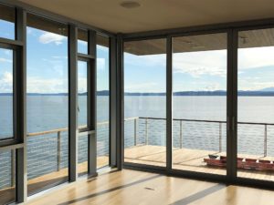 Floor-to-ceiling windows overlooking a waterfront deck and calm blue water